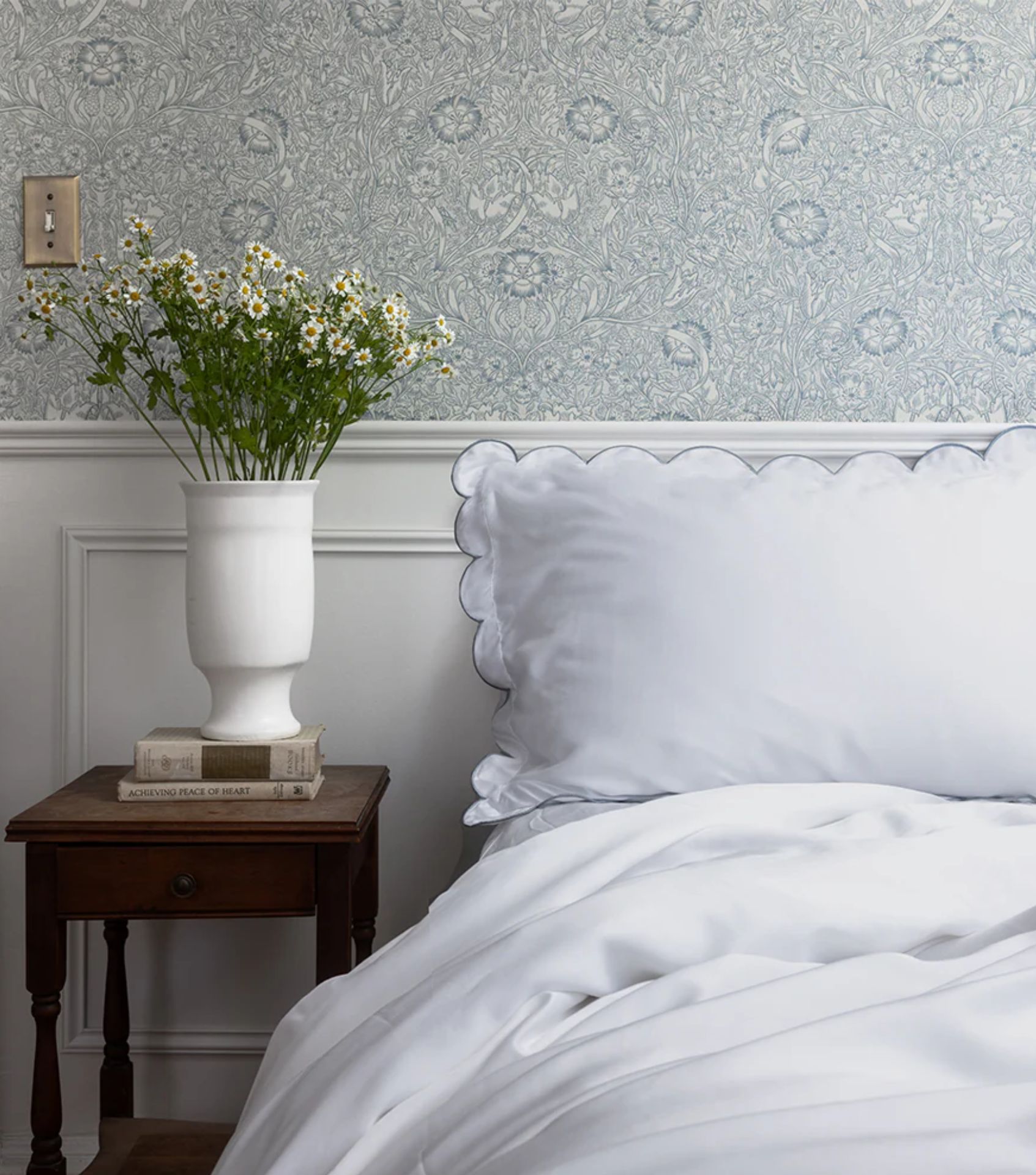 Close-up of white scalloped Eucalypso bedding made from sustainable TENCEL™ Lyocell fabric. The soft, non-toxic, and breathable sheets are made with the brand’s eco-friendly and ethical bedding design, styled with a floral wallpaper and fresh flowers.